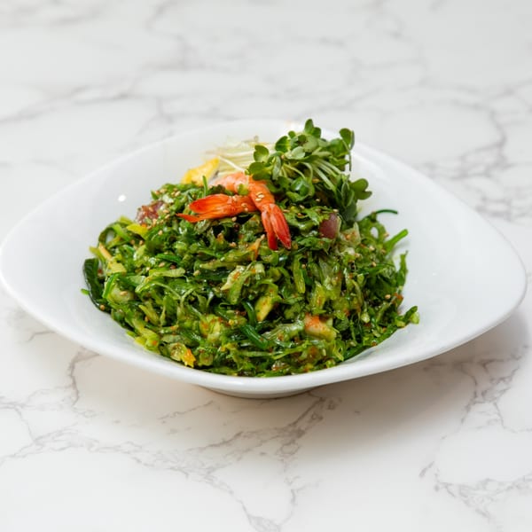 A vibrant green seaweed salad with shrimp and microgreens in a white bowl on a marble surface.