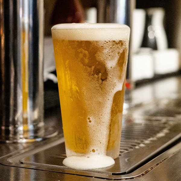 A freshly poured glass of beer with overflowing foam sits on a bar drip tray.