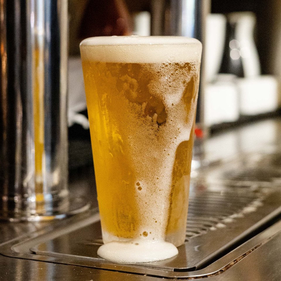 A freshly poured glass of beer with overflowing foam sits on a bar drip tray.