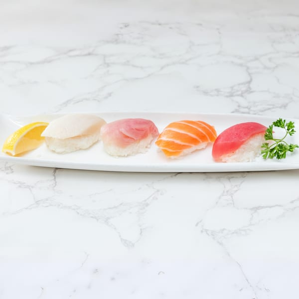 A white rectangular plate holds five pieces of nigiri sushi with a lemon wedge and parsley.