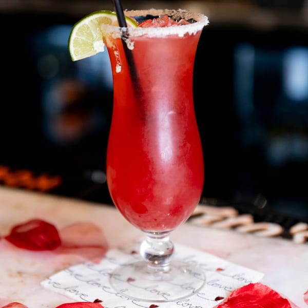 A vibrant red cocktail with a salted rim, lime wedge, and black straw, on a bar.