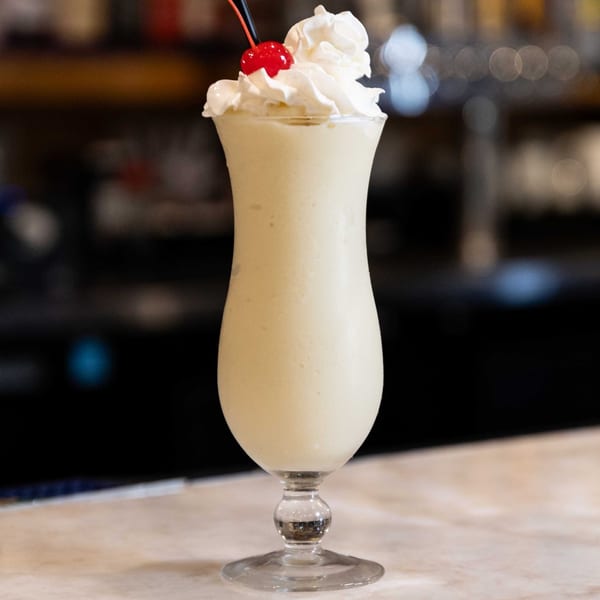 Creamy white cocktail with whipped cream, a red cherry, and a black straw in a tall glass.