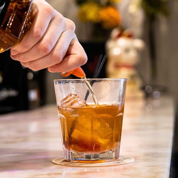 A hand pours amber liquid from a bottle into a glass with ice and a cherry on a bar.