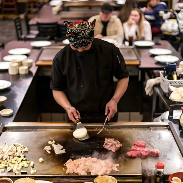 A Sakura Chaya chef cooks chicken, steak, shrimp, and vegetables on a hibachi grill for customers.