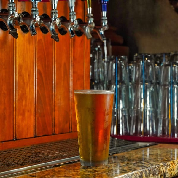 A cold beer sits on a bar counter with multiple beer taps and stacked glasses in the background.
