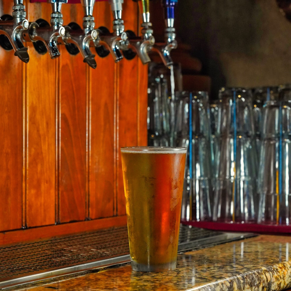 A cold beer sits on a bar counter with multiple beer taps and stacked glasses in the background.