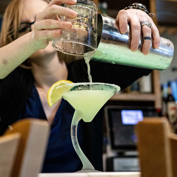 A bartender pours a green cocktail from a shaker into a martini glass with a lemon slice.