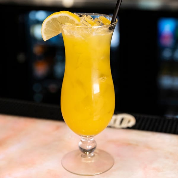 A vibrant yellow cocktail with ice, a lemon slice, and a black straw on a bar.
