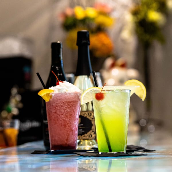 Two colorful cocktails, one red and one green, garnished with fruit and cherries, on a bar.