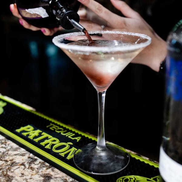 A bartender pours a dark liquid into a sugar-rimmed martini glass with a red cherry inside.