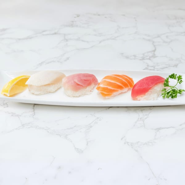 A white rectangular plate holds four pieces of nigiri sushi, a lemon wedge, and parsley on marble.